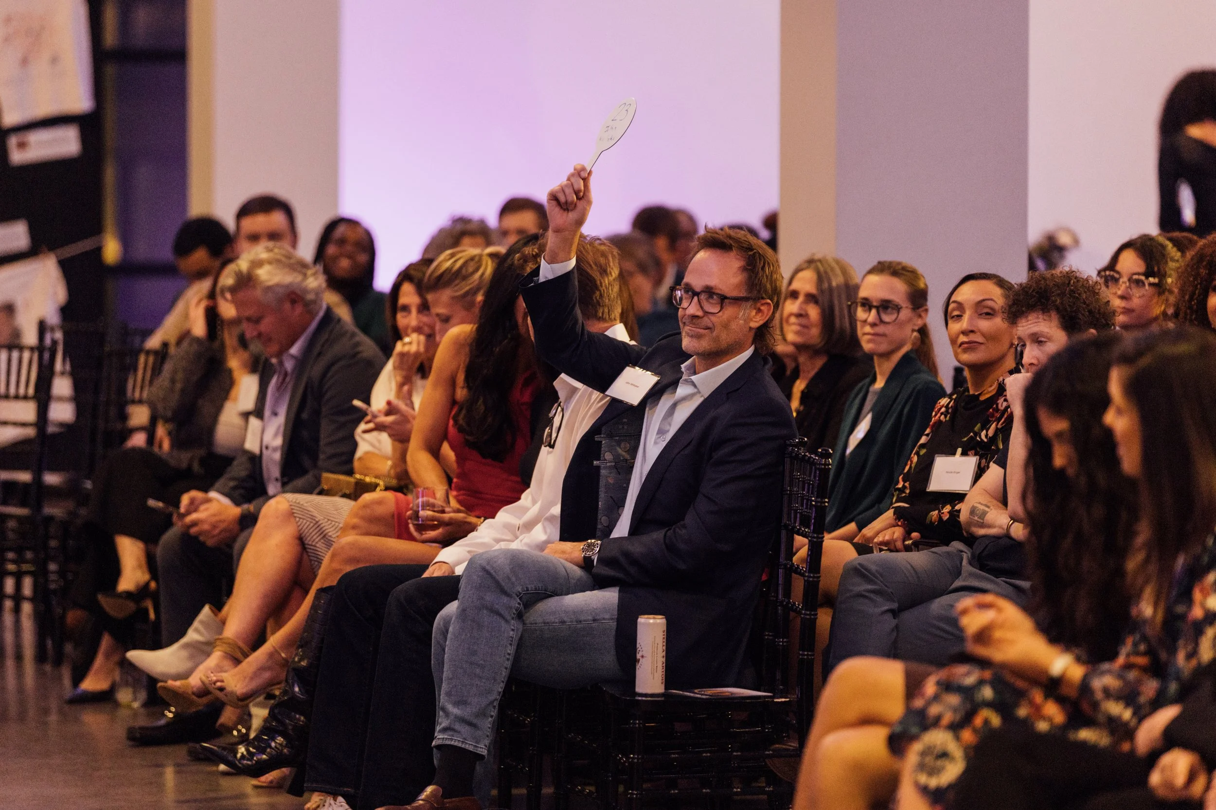 A man in a suit raising his hand while holding a paper paddle at a conference, surrounded by seated attendees in a dimly lit room.