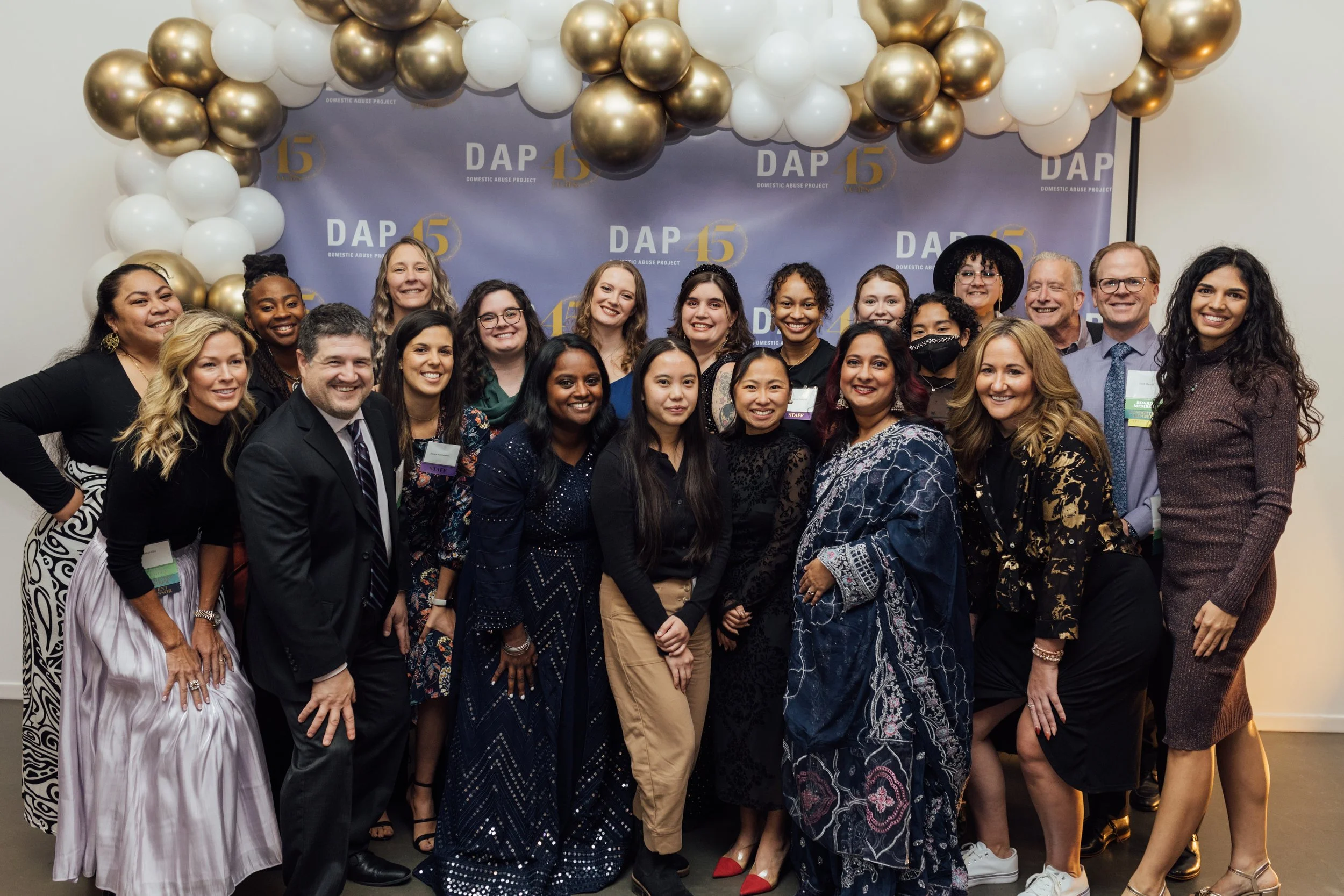 Group photo of diverse people at an event celebrating the 15th anniversary of the Domestic Abuse Project, with a backdrop decorated with balloons and the organization's logo.