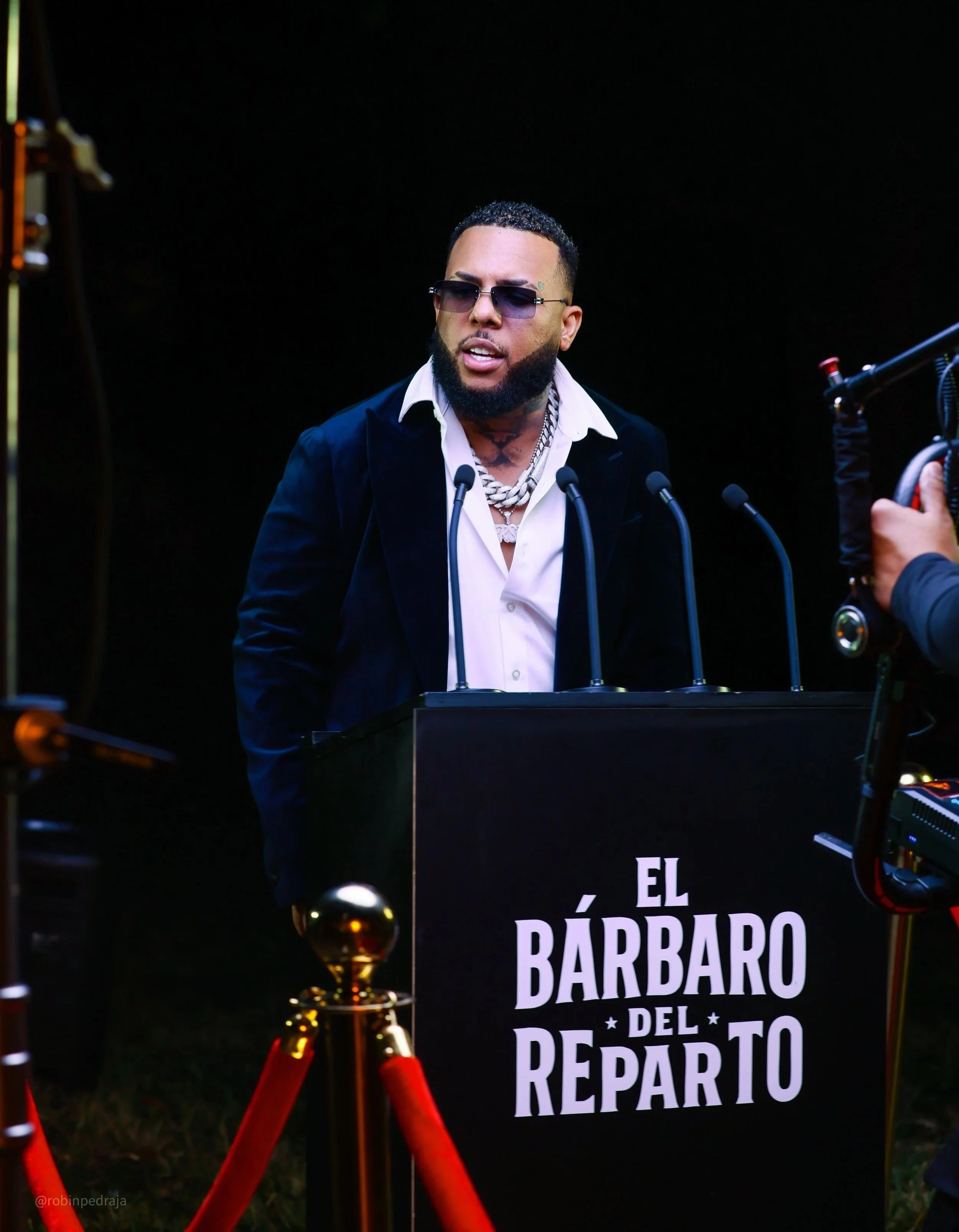 Wow Popy, hombre con gafas oscuras, barba y collar de cadena, de pie detrás de un podio que dice 'El Bárbaro del Reparto'.