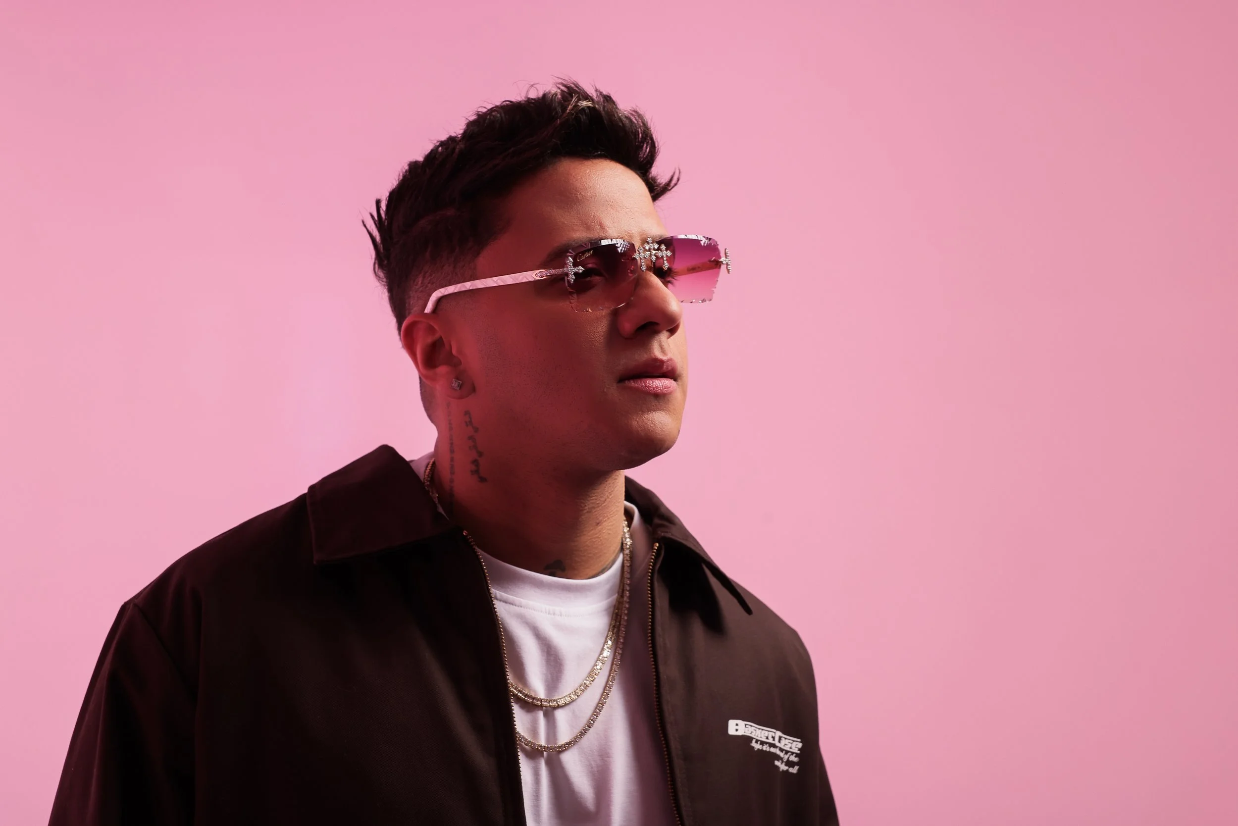 Divan con gafas rosas, peinado estilo moderno, roupa negra con cadenas, fondo rosa.