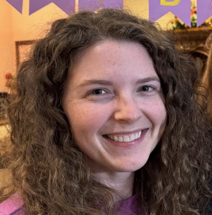A smiling woman with long, curly brown hair, wearing a pink top, with a colorful background behind her.