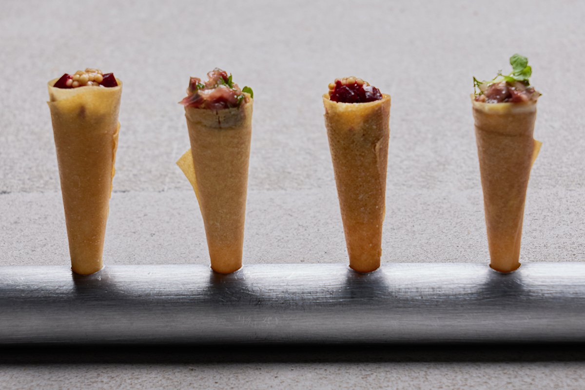 Four small cone-shaped pastries filled with various ingredients, standing on a metallic holder against a neutral background.
