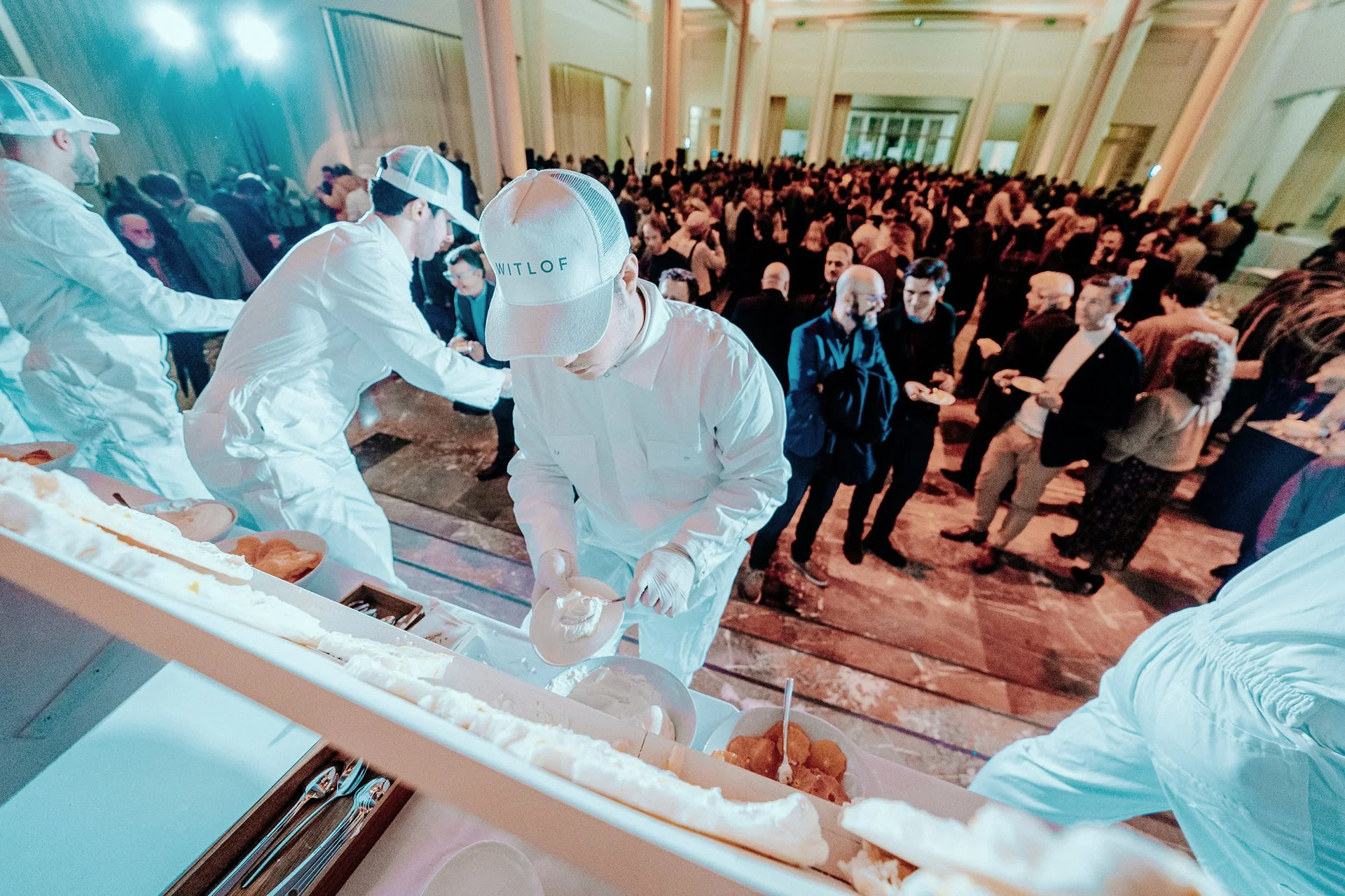 People in white uniforms and hats serving ice cream at a buffet table during a large event in a hall filled with many attendees.