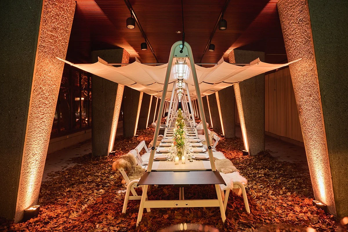 Long dinner table set for a formal event with flowers and candles, under a canopy in a decorated outdoor area with leaf-covered ground.
