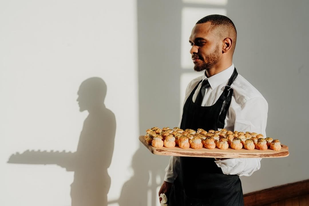 A waiter holding a tray of small pastries in front of a plain wall with sunlight casting a shadow of a person playing a keyboard.