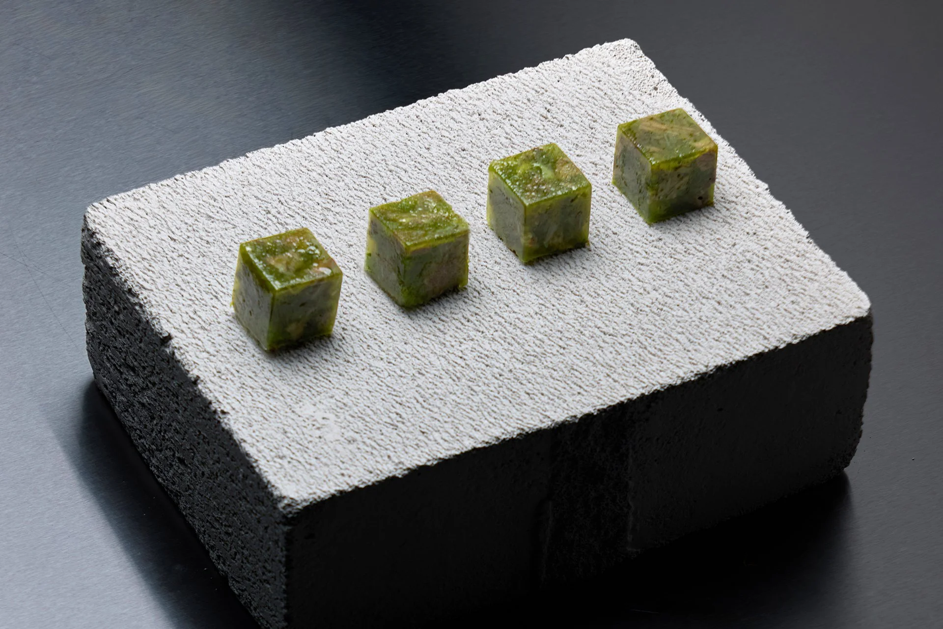 Four small cube-shaped desserts with green and white marbled appearance placed on a textured white surface.
