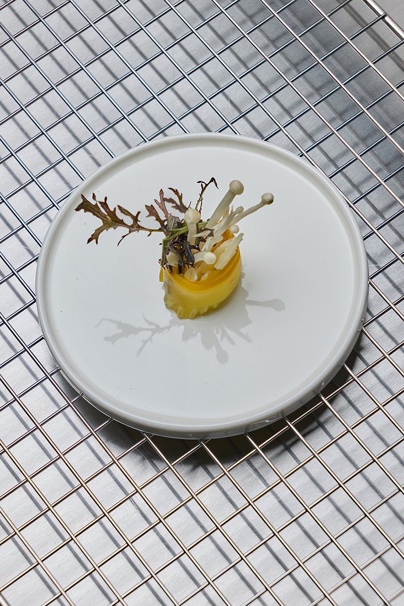 A white plate with a small bundle of white enoki mushrooms, a yellow pickled vegetable, and sprigs of decorative leaves on a wire rack.