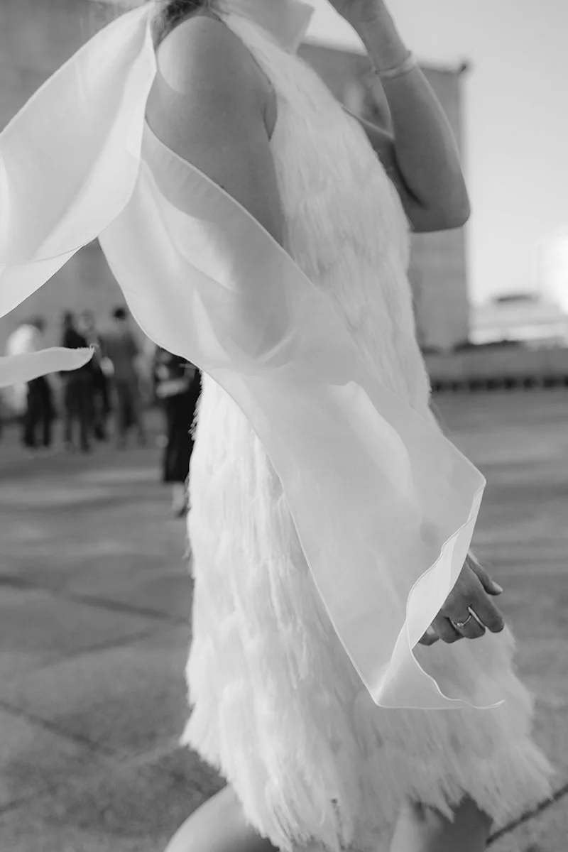 A woman in a light, textured dress with sheer, flowing fabric draped over her shoulder, standing in an outdoor area with people in the background.