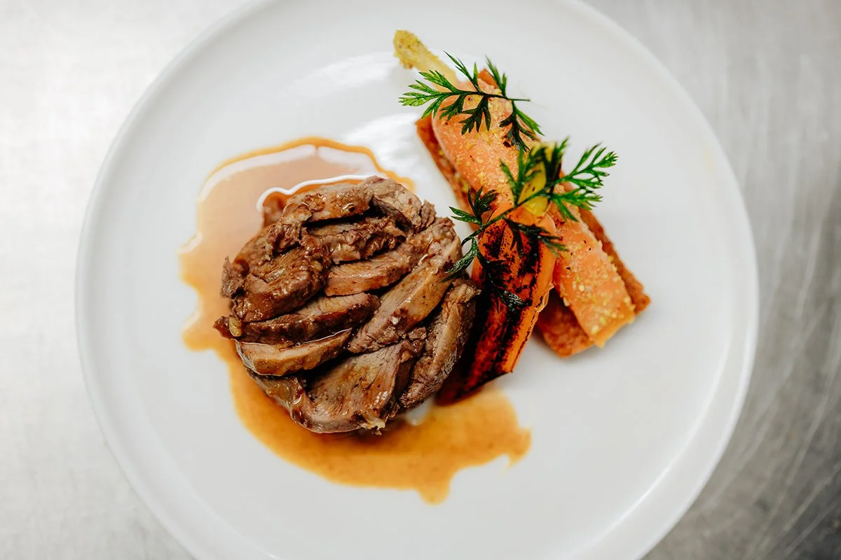 A plated dish with slices of cooked meat, roasted carrots garnished with fresh herbs, and a sauce on a white plate.