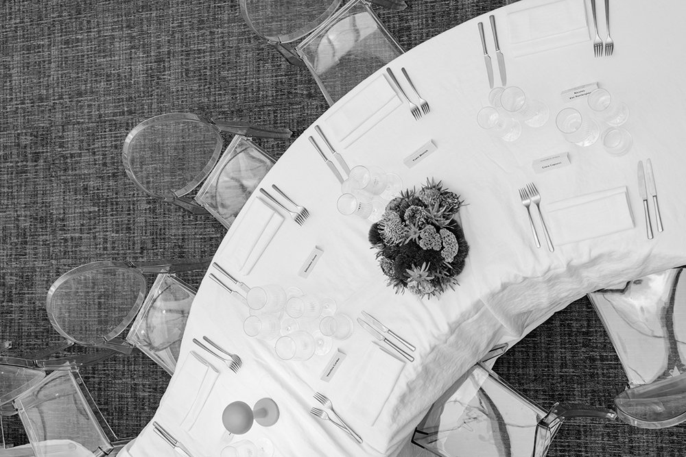 An overhead view of a round banquet table with a white tablecloth, set with silverware, glasses, and name cards, with a floral centerpiece, surrounded by clear acrylic chairs.