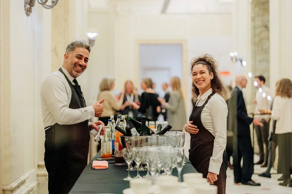 Two smiling bartenders, a man and a woman, standing behind a drinks station at a social event. The man is giving a thumbs-up, the woman has her hand on her stomach. There are bottles, glasses, and drinks on the table, with a group of people socializi