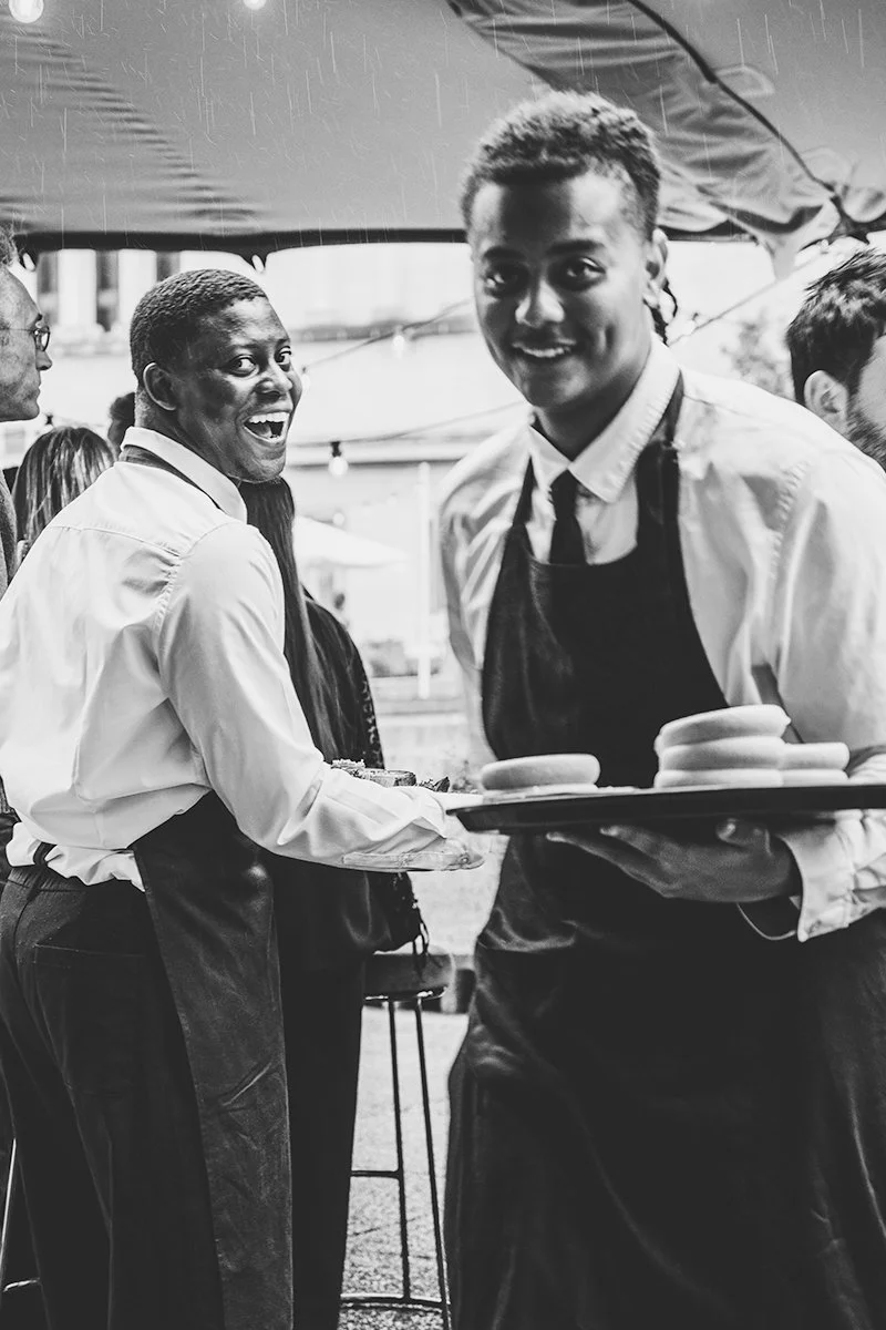 People serving food at an outdoor event, smiling and engaging with attendees, under a tent.