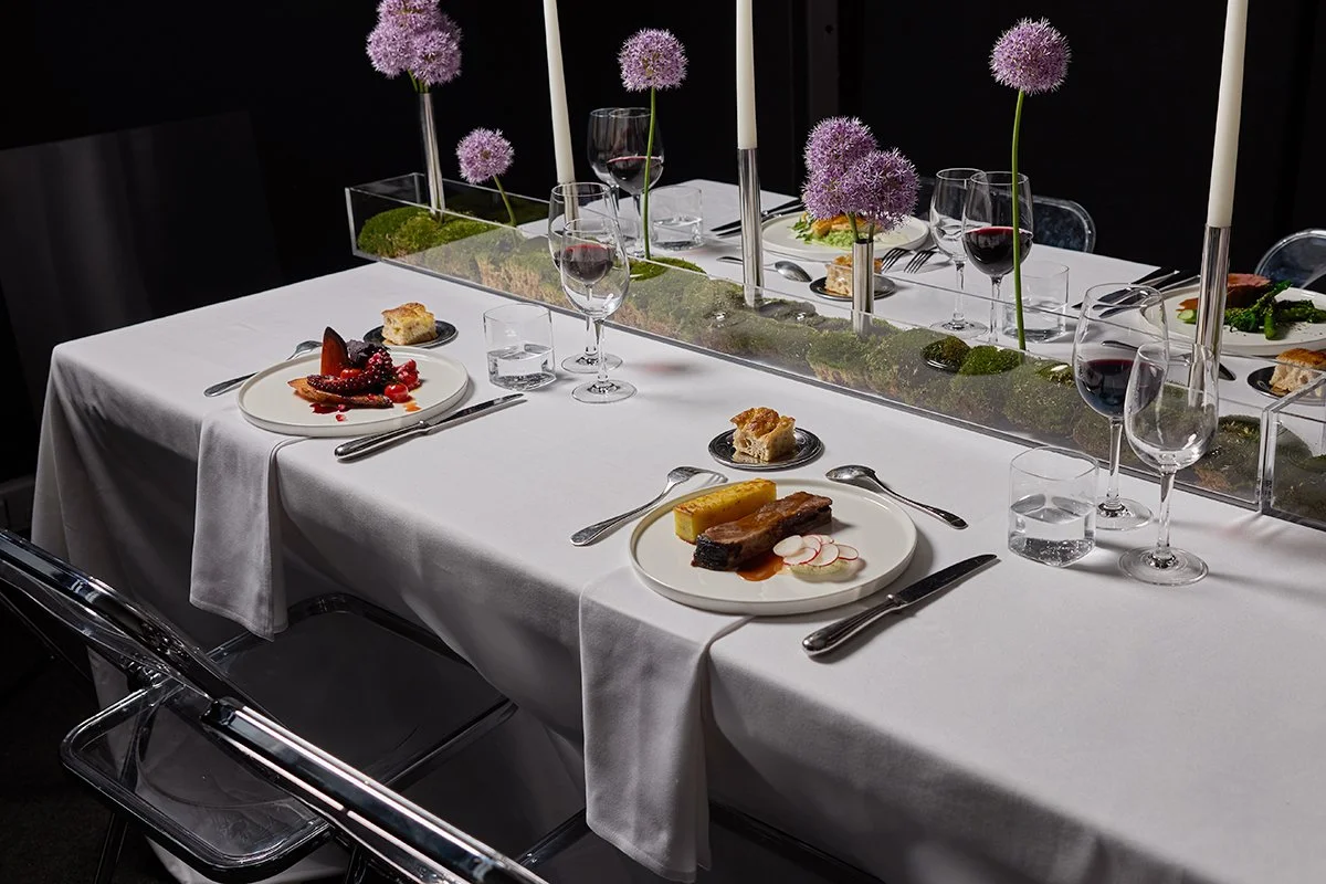 Elegant dining table with white tablecloth, decorated with purple flowers in tall vases, set with plates of gourmet food, glasses of red wine, and candles.