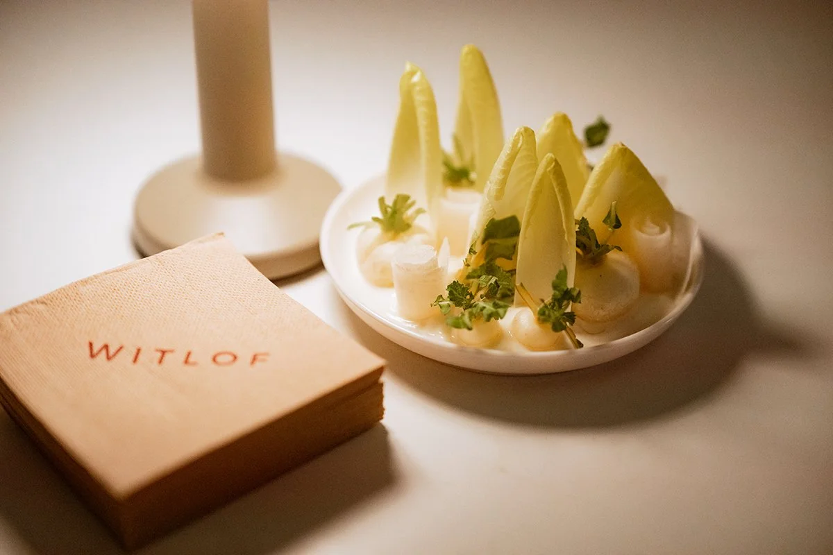 A plate of sliced artichokes garnished with herbs and onions, a stack of WITLOF-branded napkins, and a candle on a white table.
