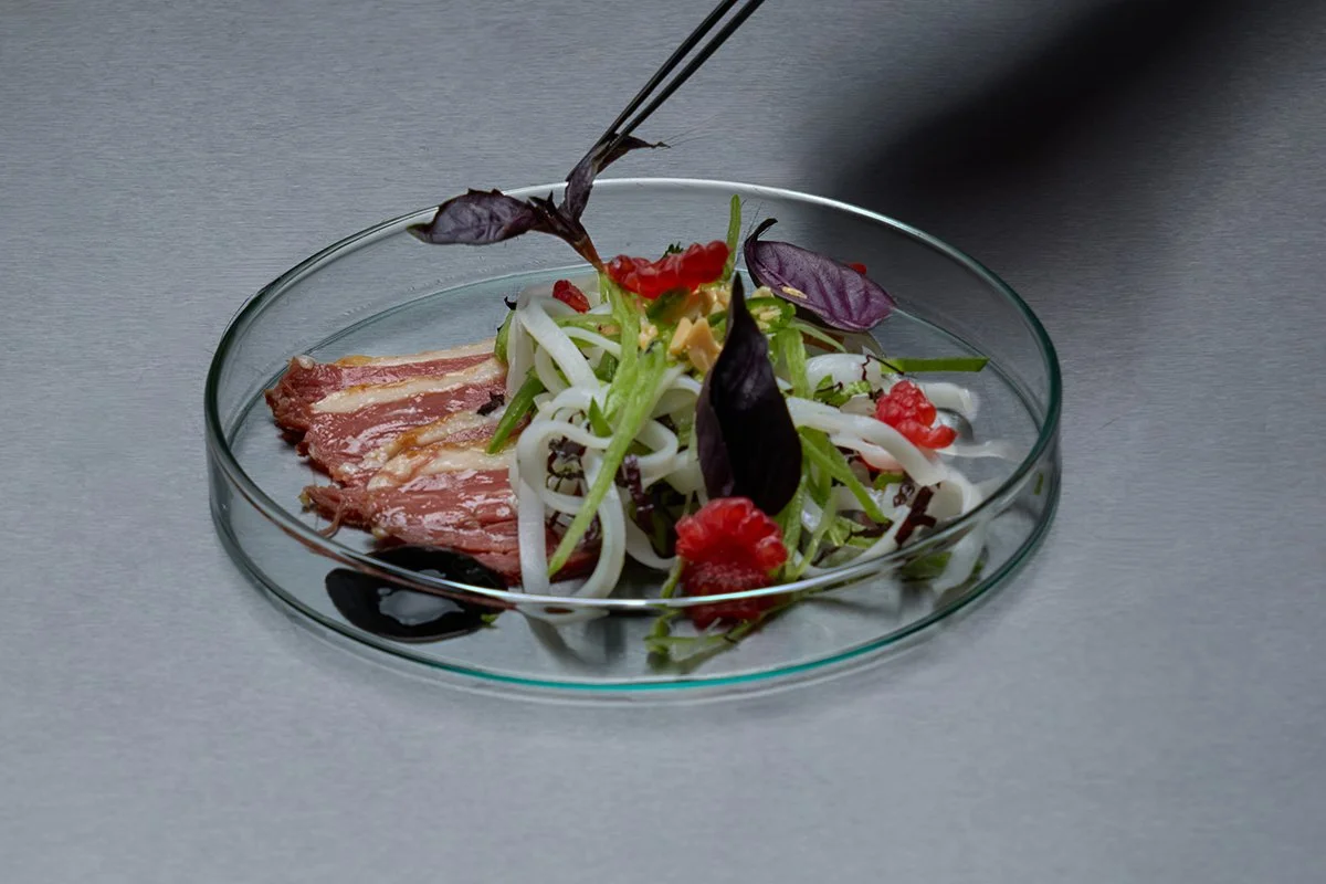 A glass dish containing a salad with sliced meat, mixed greens, herbs, and red berries.