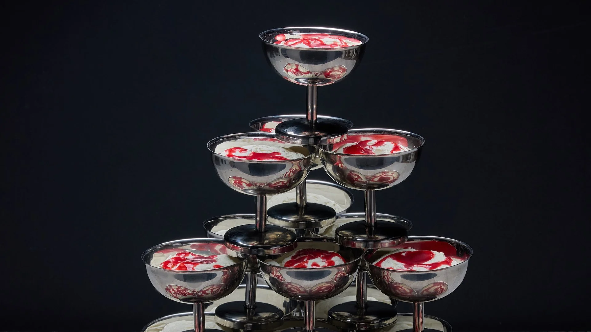 A pyramid stack of martini glasses filled with a creamy dessert topped with red syrup on a black background.