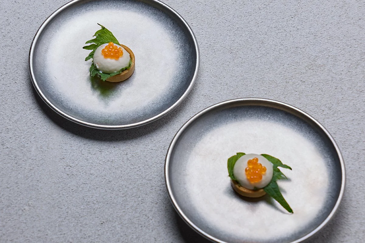 Two small plates each with a bite-sized appetizer consisting of a mound of white soft cheese or cream with orange fish eggs on top, garnished with green leaves, on a gray textured surface.