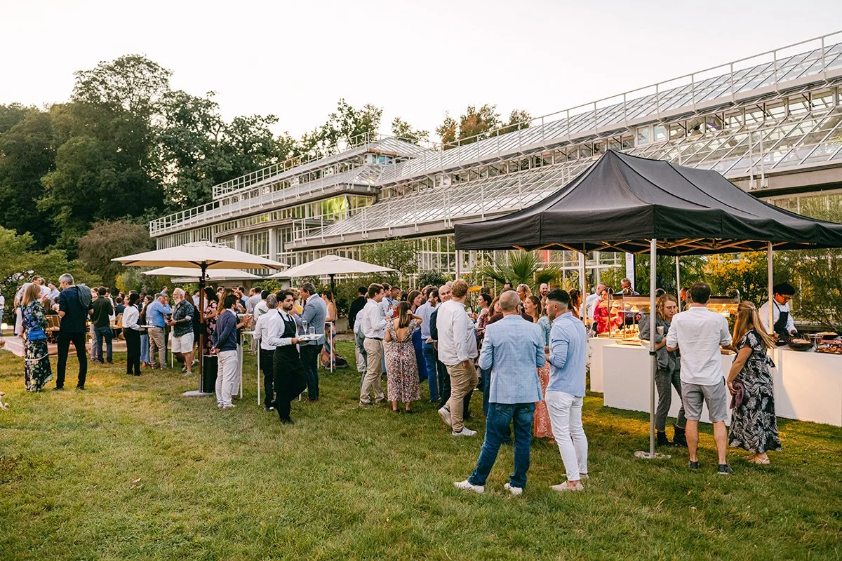 Outdoor social gathering with people chatting and enjoying food under umbrellas and a canopy outside a glass greenhouse or conservatory building in the evening.