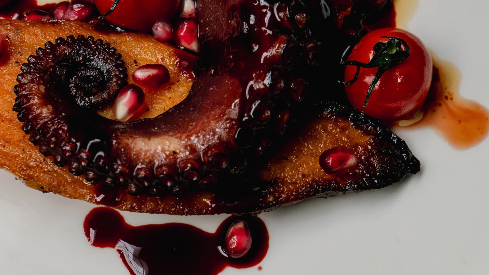 A cooked octopus tentacle garnished with pomegranate seeds and cherry tomatoes, drizzled with a dark sauce, on a white plate.