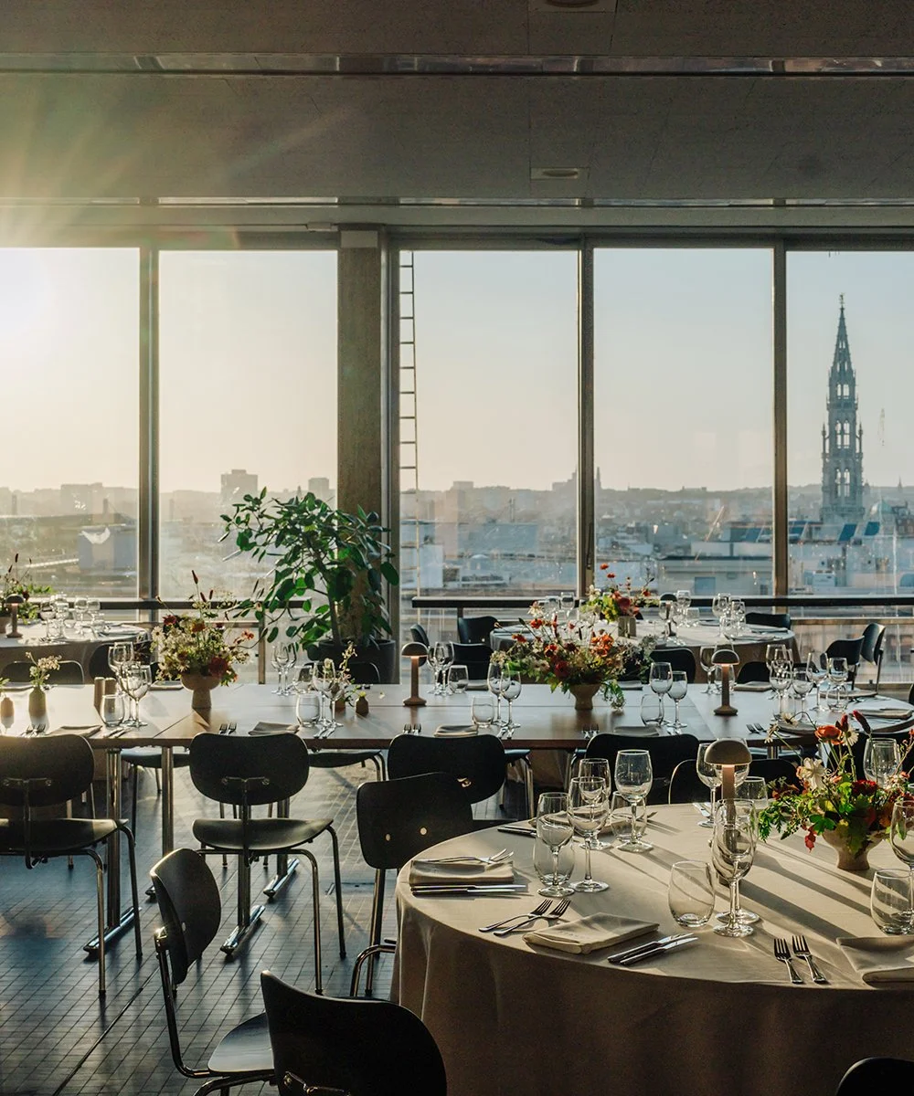 Elegant dining room with large windows, decorated with flower arrangements, set for a formal event.