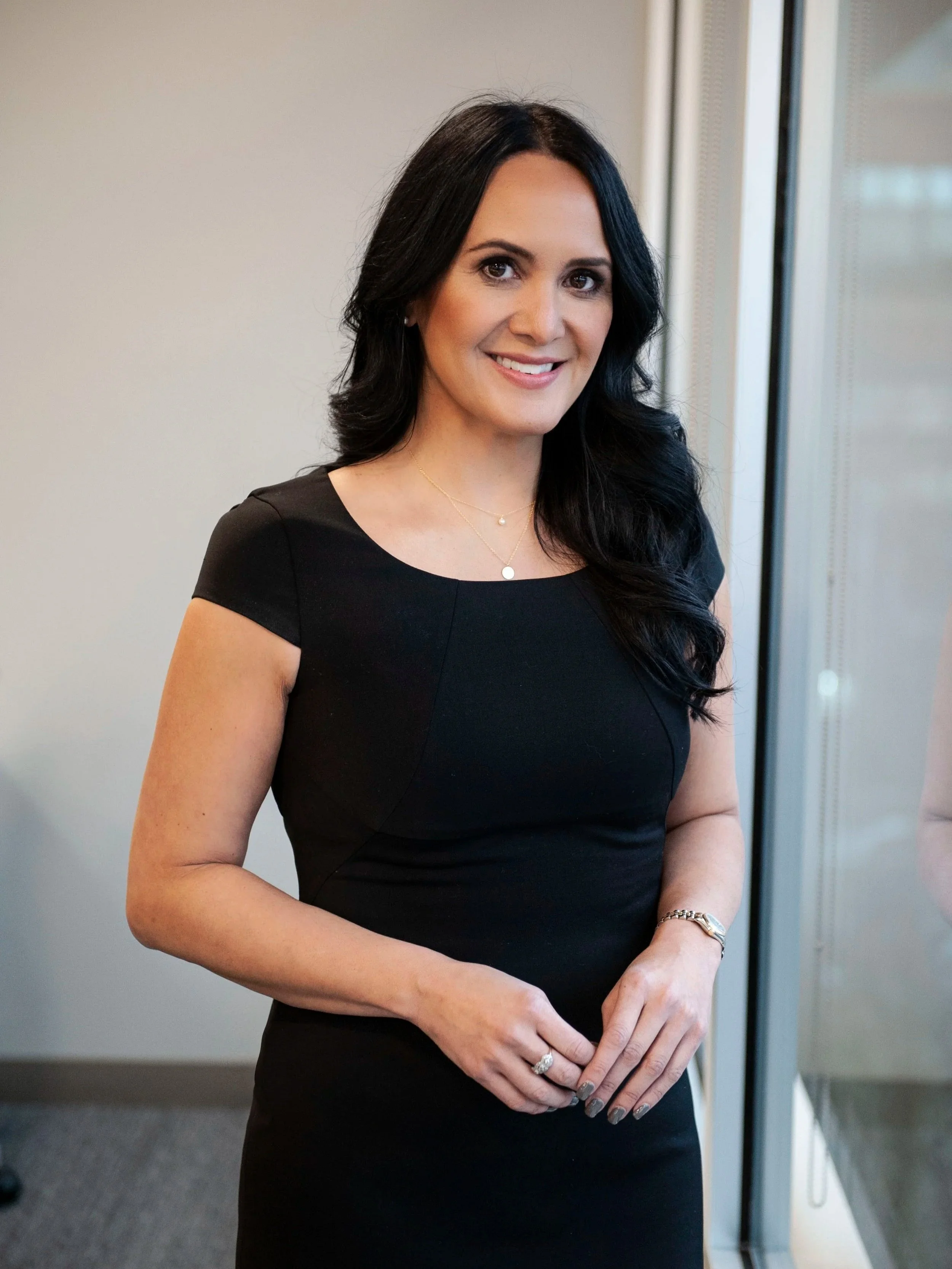 Wendy Estela. Estela Law. Legal Expertise in Connecticut, New York, and Utah. A woman with long dark hair wearing a black dress and jewelry, smiling and standing indoors near a window.
