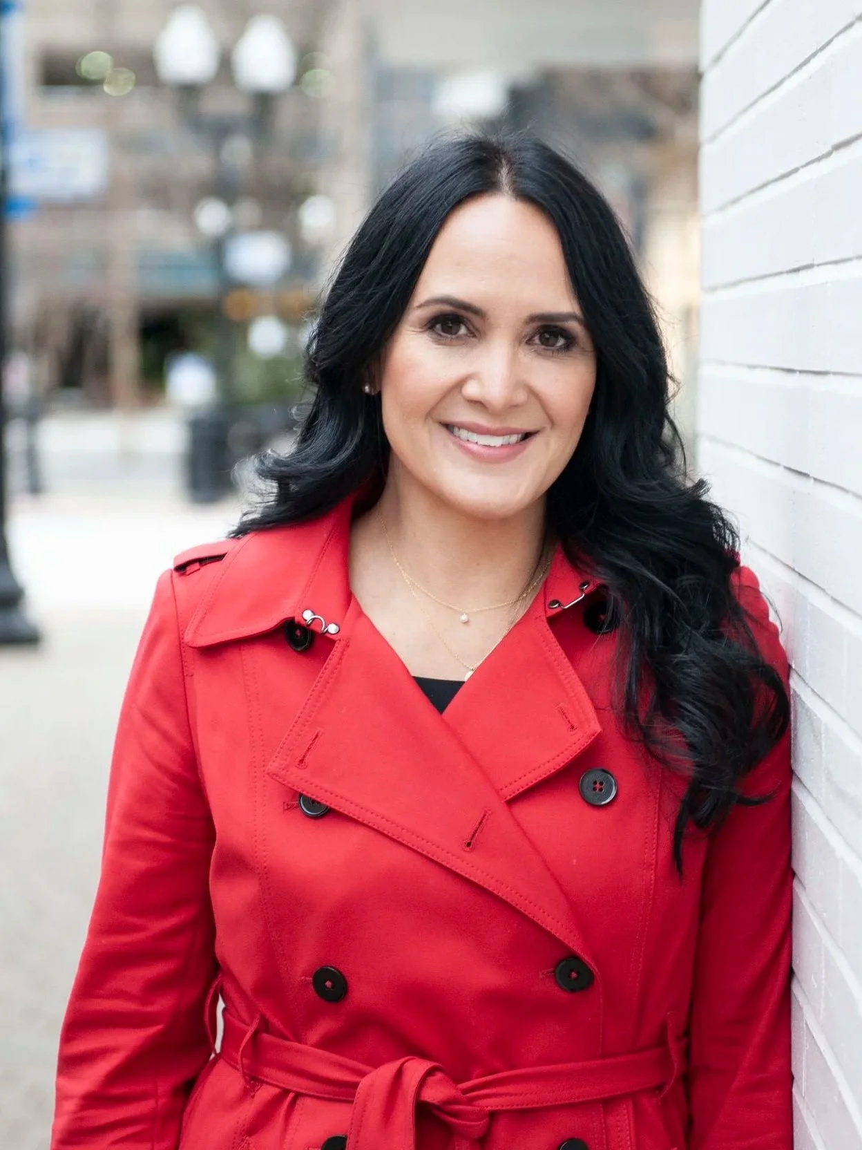 Wendy Estela. Estela Law. Legal Expertise in Connecticut, New York, and Utah. A woman with long black hair, smiling, wearing a red trench coat, standing outdoors next to a white brick wall.