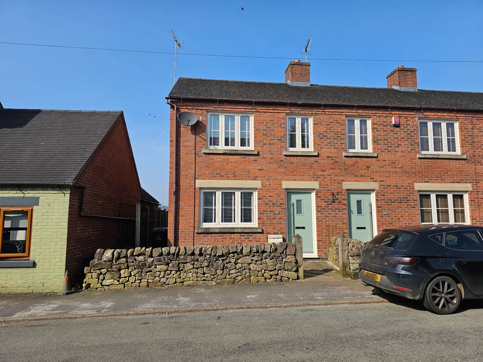 Front elevation of a beautifully presented red brick two bedroom end town house on High Street Dilhorne with green doors, stone wall frontage and parked car