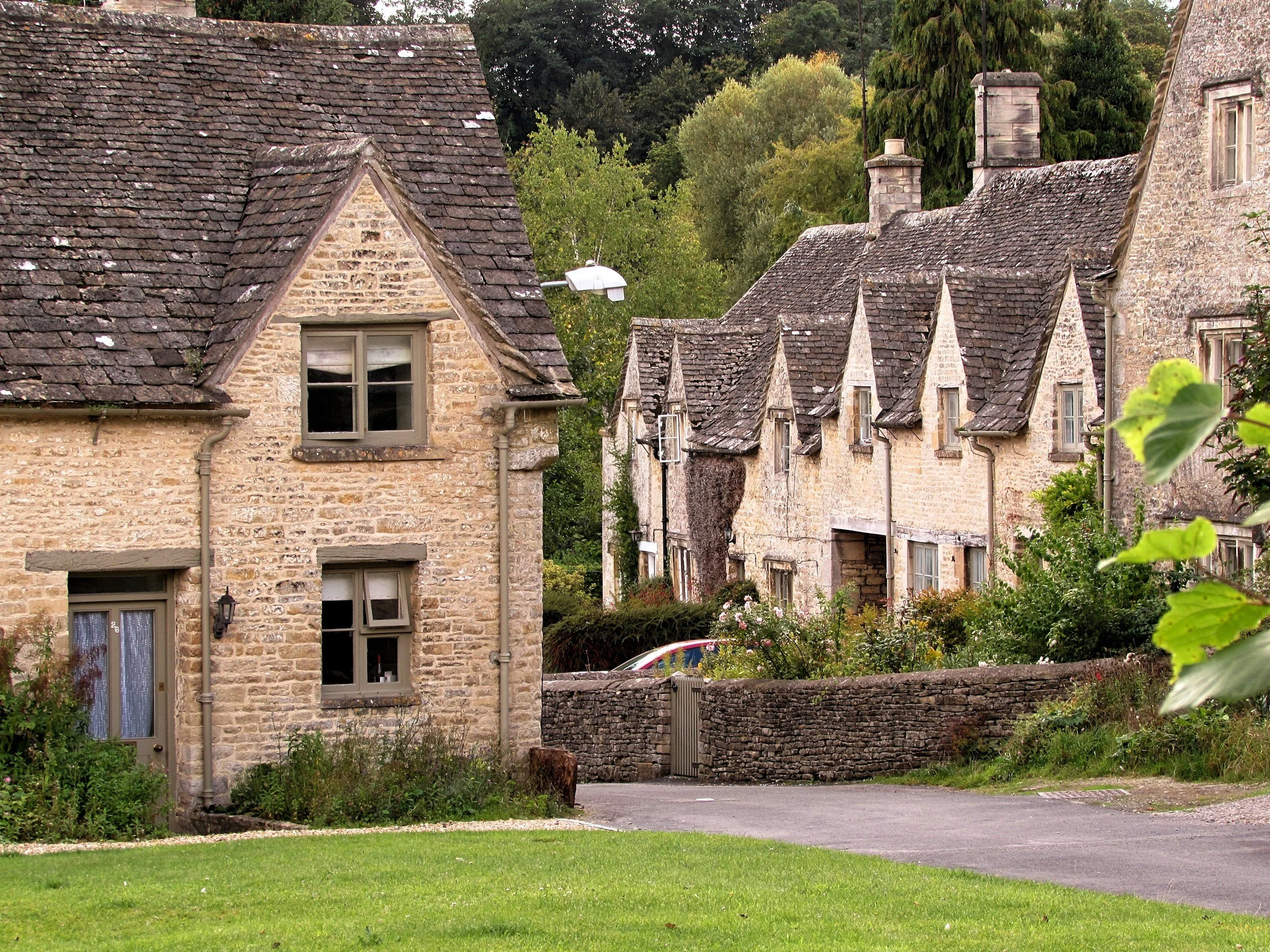 A view of old stone houses in Cotswold with sloped roofs and small windows, surrounded by greenery and a stone wall, in a rural setting.
