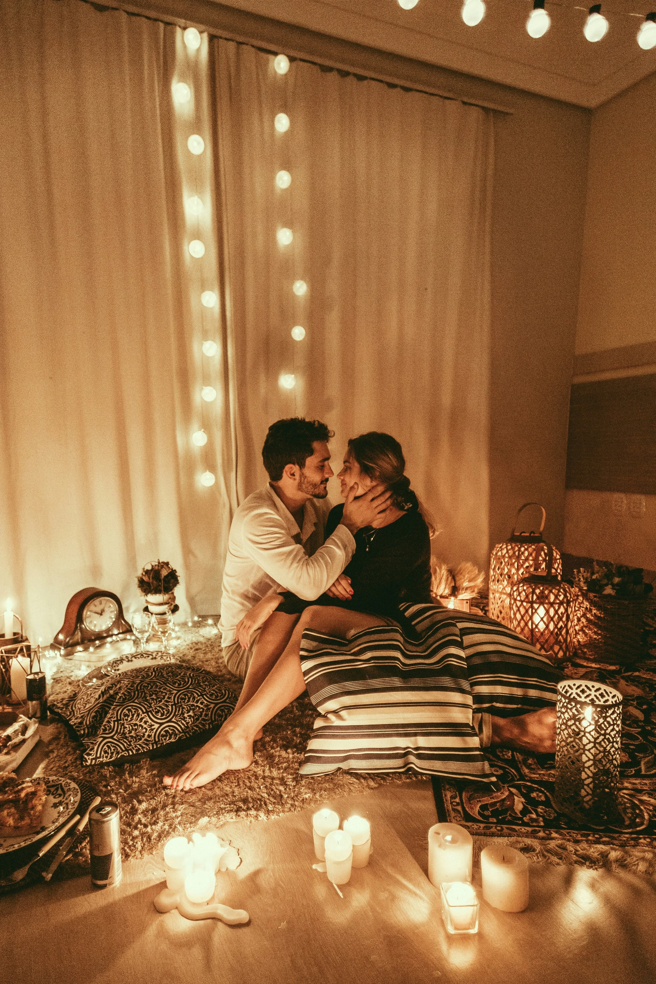 A couple sitting closely together on the floor in a dimly lit room decorated with candles, string lights, and lanterns, sharing an intimate moment.