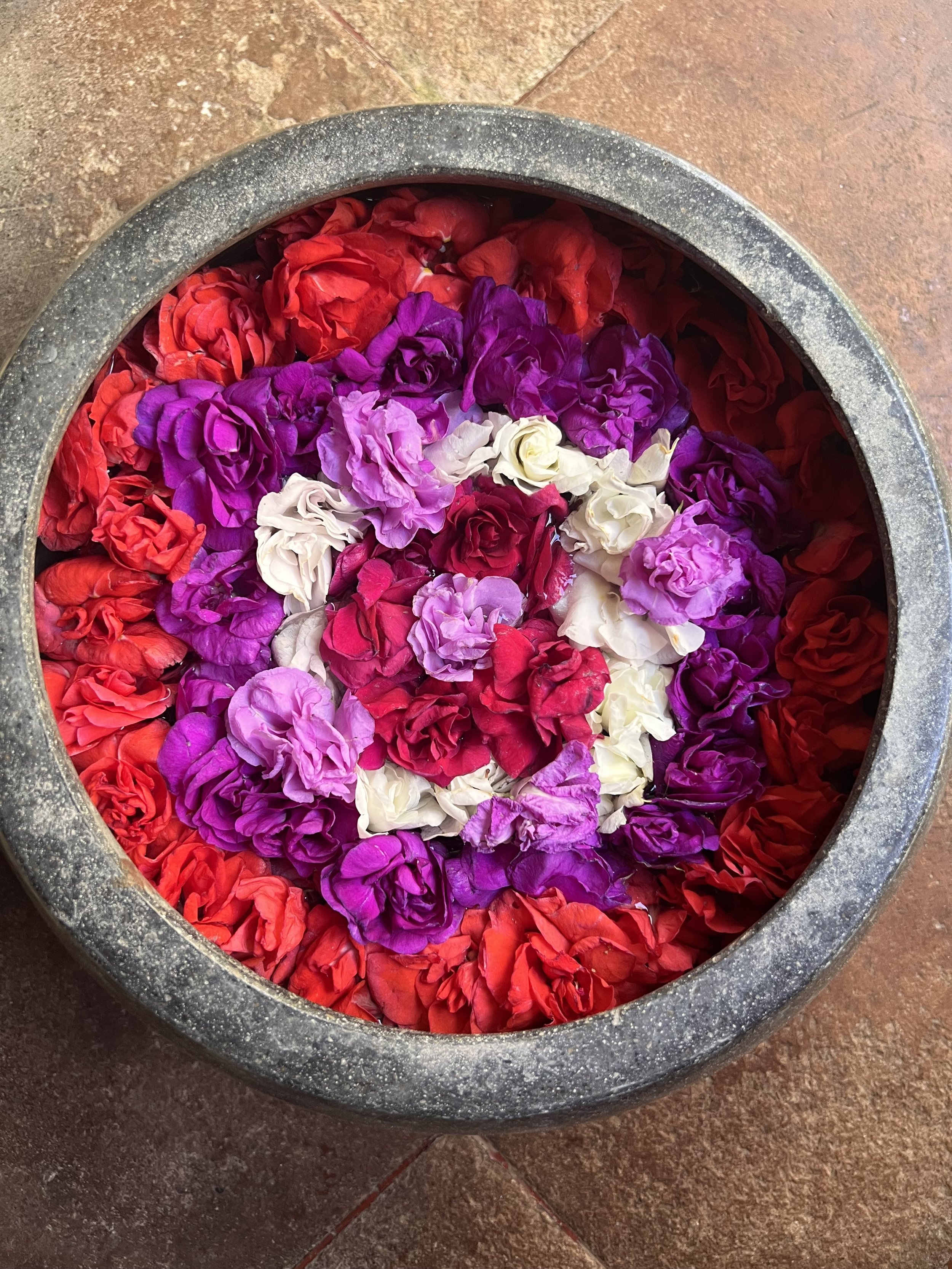 A circular stone bowl filled with colorful flower petals, including red, purple, white, and pink, on a tiled floor.