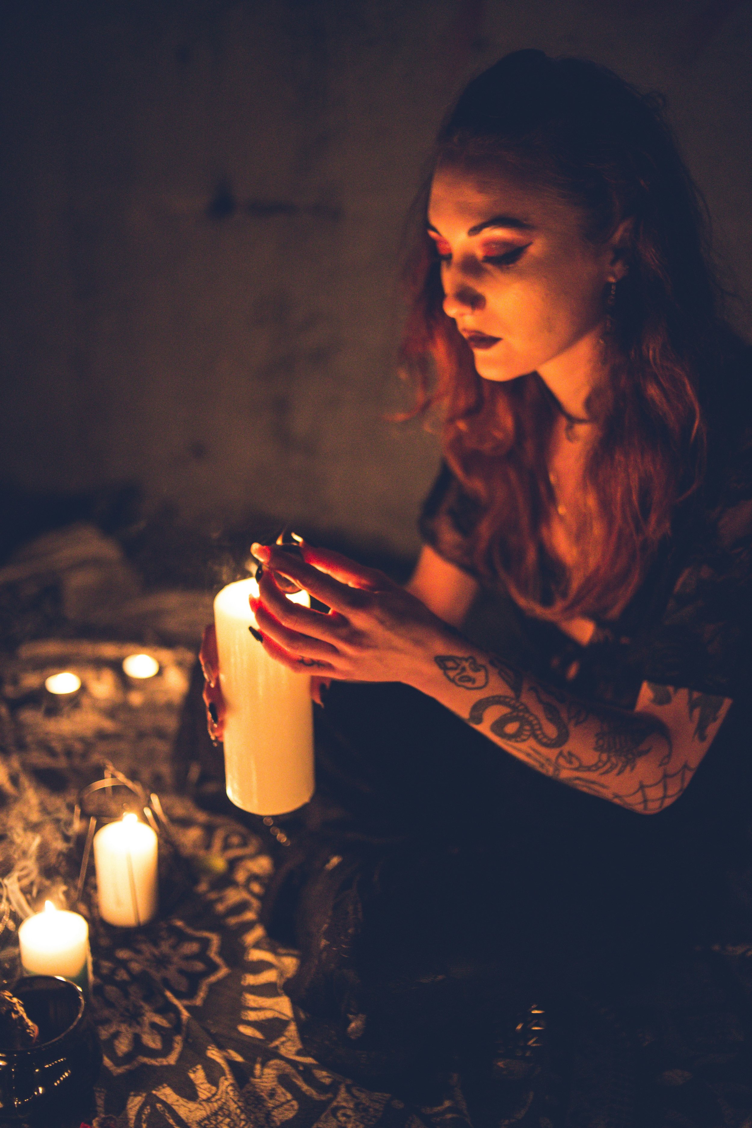 A woman with tattoos on her arm, holding a lit candle in a dark room with additional candles around her.