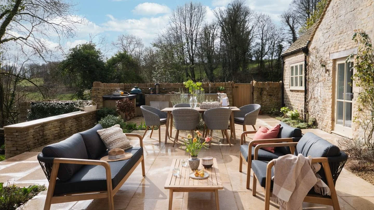 Outdoor patio in Cotswold with seating and dining area, including a sofa, chairs, a table with wine and flowers, and a barbecue grill in the background.