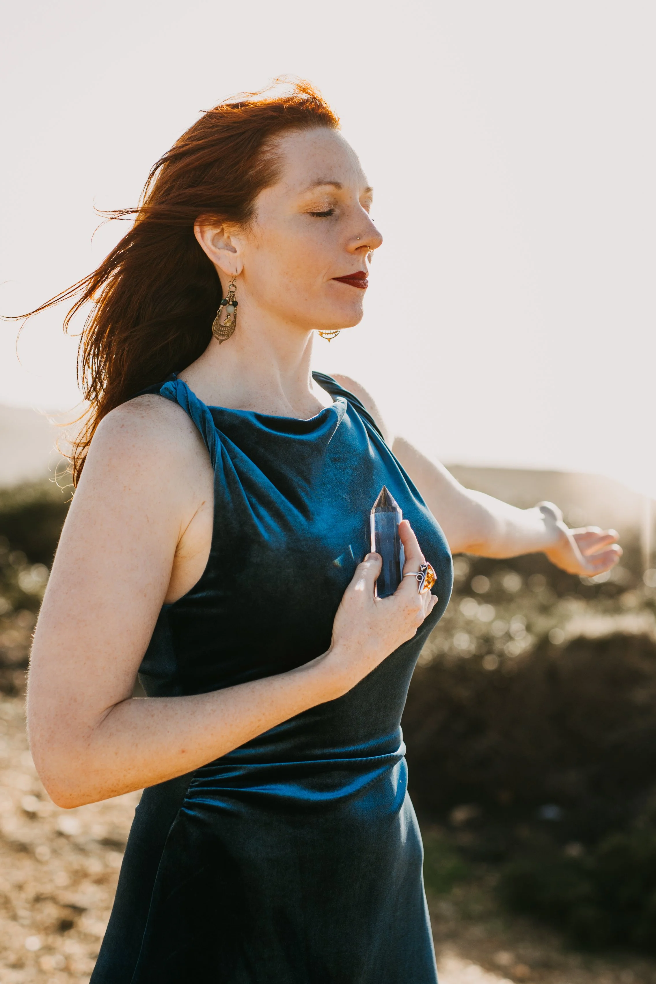 A woman with red hair and freckles practicing yoga outdoors, holding a crystal in one hand with closed eyes during sunset.