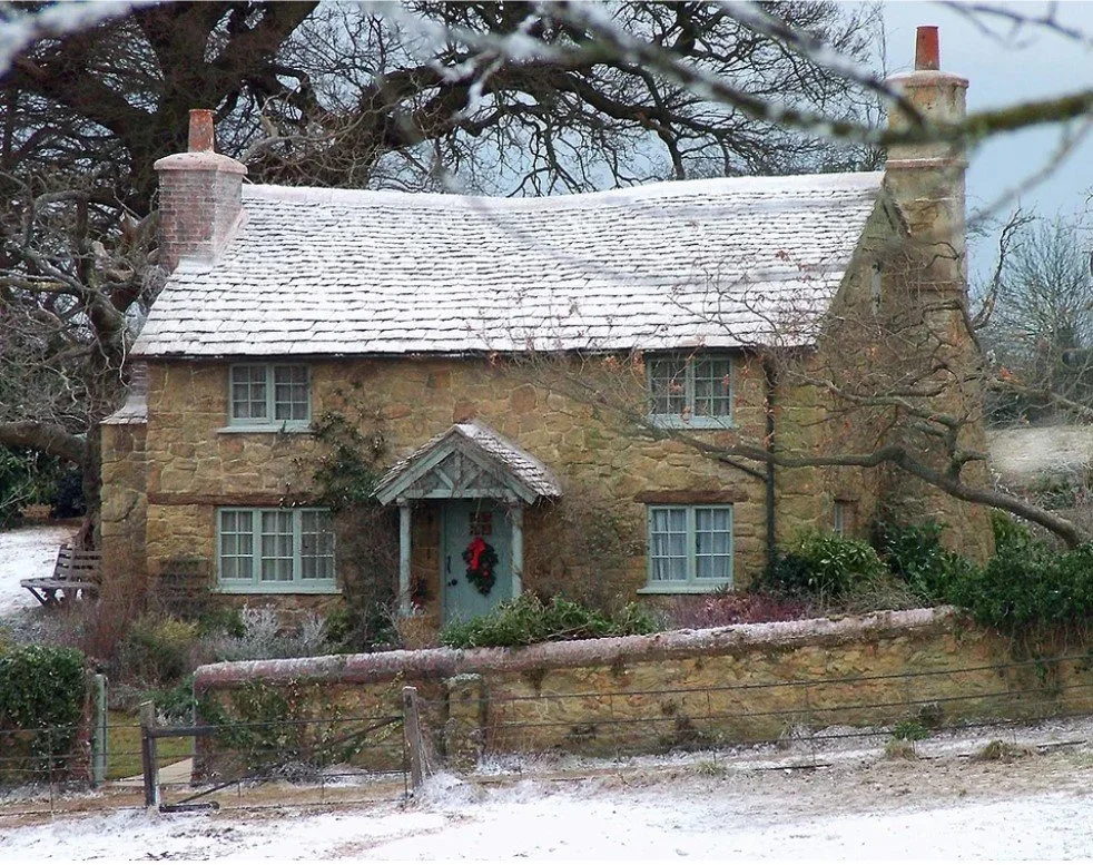 A historic stone house  in Cotswold with a snow-covered roof, two chimneys, and a wreath on the front door, set in a winter landscape with bare trees and snow on the ground.