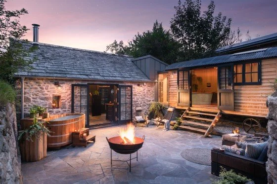 Outdoor patio area next to a stone house and a wooden cabin in Cotswold, with a fire pit, hot tub, seating, and decorative plants at sunset.