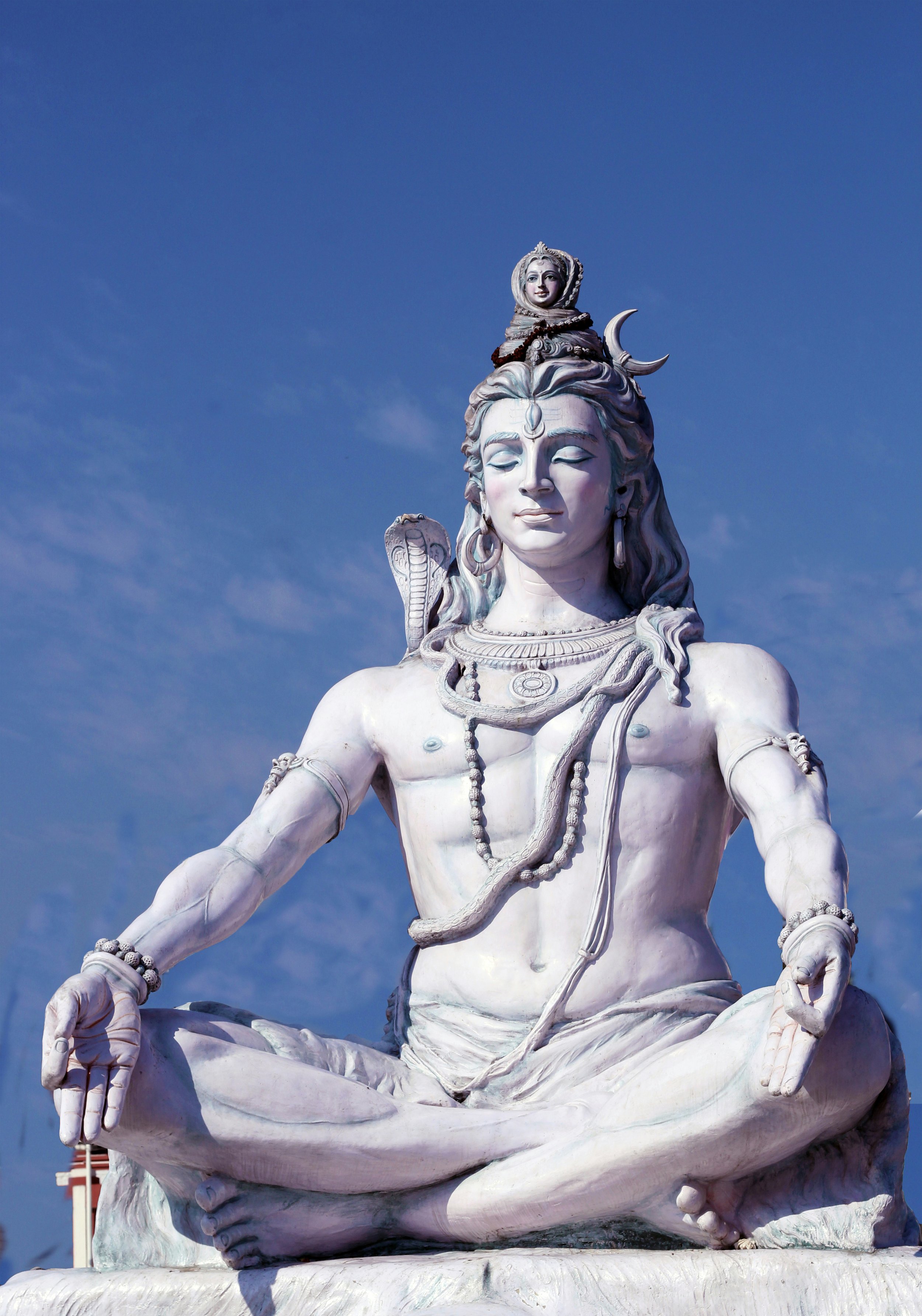 White marble statue of Lord Shiva in a seated meditative pose against a blue sky with some clouds.