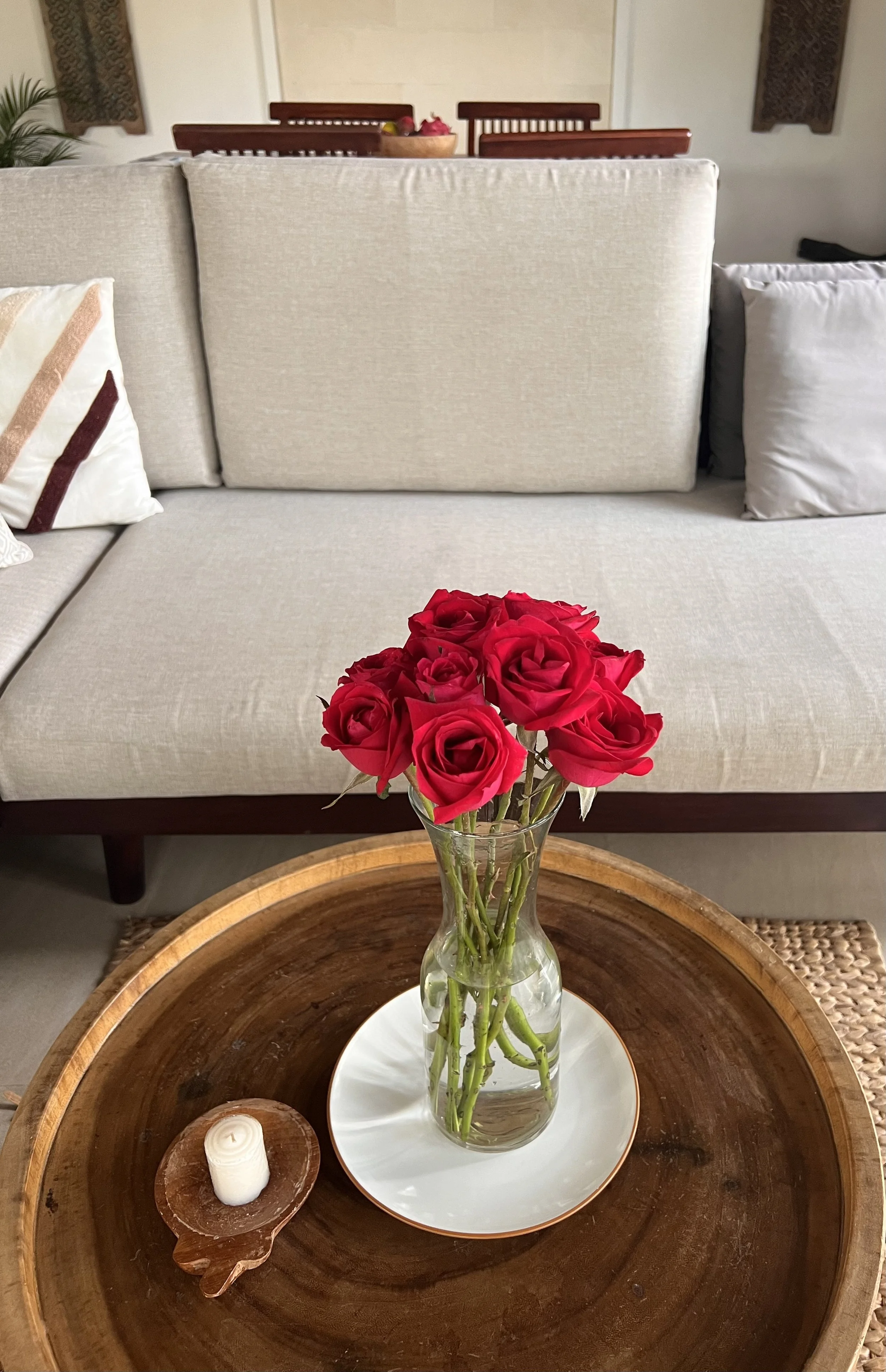 A living room with a beige sofa, decorative pillows, and a wooden coffee table. On the table is a glass vase with red roses and a white plate underneath, along with a small white candle on a wooden holder.