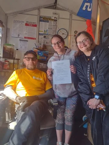 Three people inside a work or medical facility. One man seated in a wheelchair wearing a yellow shirt and glasses, two women standing nearby holding a document. The background shows posters, a clock, and equipment.