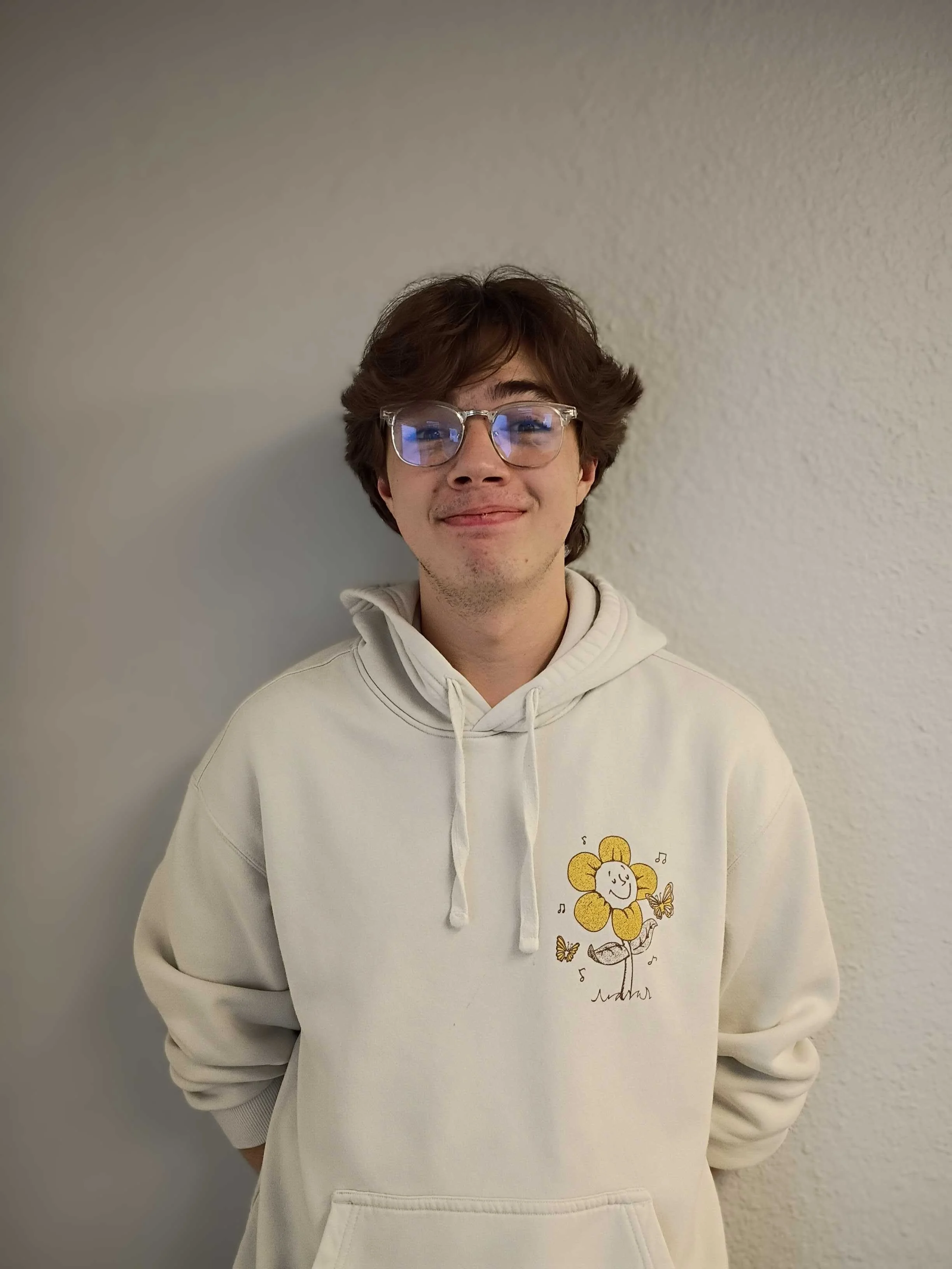 A young man with brown hair and glasses smiling while wearing a white hoodie with a flower cartoon design on the front. He stands against a light-colored textured wall.
