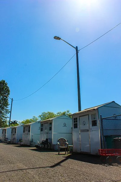 A row of white mobile homes or trailers with a tall streetlamp and utility wires against a clear blue sky.