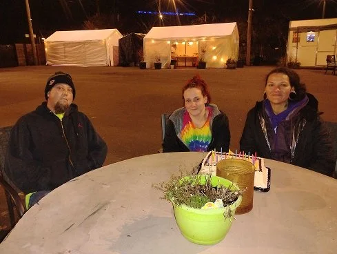 Three people sitting around a round table with a birthday cake featuring candles and a green plant in a yellow pot, outside at night with tents in the background.