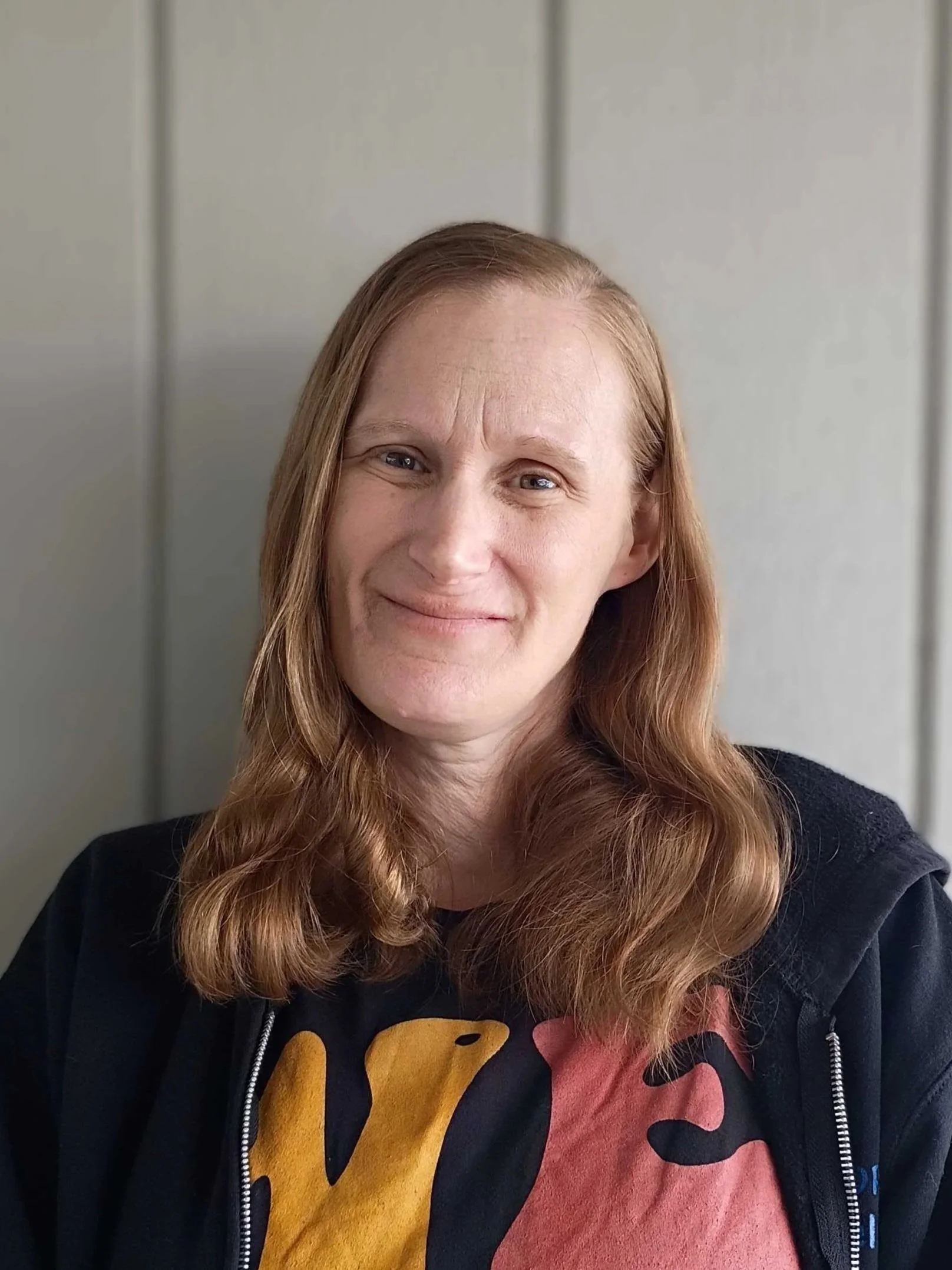 A woman with long, wavy red hair smiling against a plain, light-colored wall.