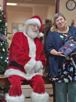 Santa Claus sitting next to a woman holding a wrapped gift, in a decorated room with a Christmas tree.
