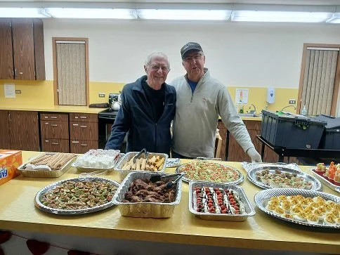 Two men standing behind a table with various casseroles and appetizers in a room with wooden cabinets and yellow walls.