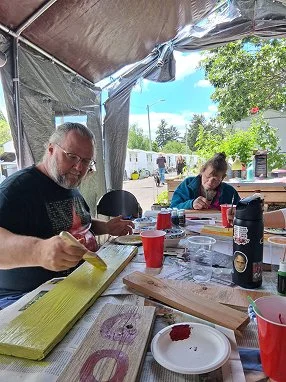 Two men sitting at a table outdoors under a canopy, painting on canvases. Various painting supplies and drinks are on the table, with trees and a trailer visible in the background.