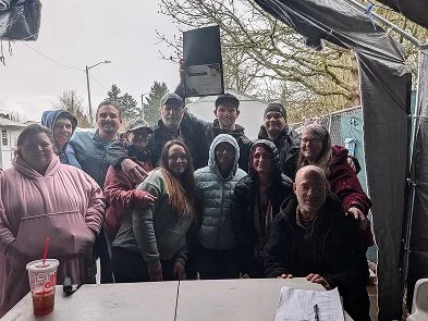 Group of people outdoors under a tent, standing for a photo, some wearing jackets and hoodies, with a drink on the table in front.