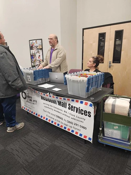 A table with a sign for Neighborhood Mail Services in a community center, with two bins of mail and a woman sitting behind the table, while a man is talking to a man standing in front.