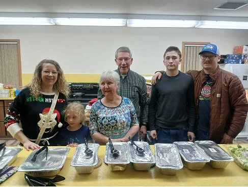 Group of six people, including children and adults, standing behind a table with covered trays, in a room with fluorescent lighting and light-colored walls, possibly at a community or social gathering.