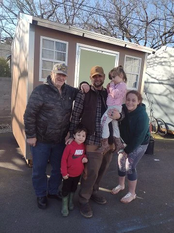 Group of five people, two men, two women, and a young girl, standing in front of a small shed or tiny house, outdoors on a paved area with trees in the background.