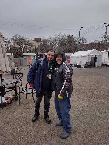 Two men standing outdoors in a parking lot with tents and small buildings behind them on a cloudy day.